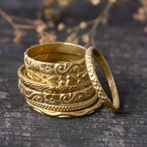 May include: A stack of six gold-toned rings with intricate designs. The rings are stacked on a dark wooden surface.