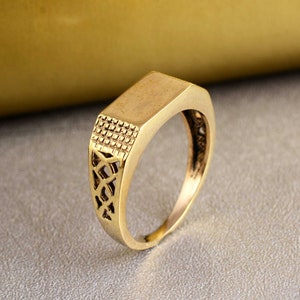 May include: A gold-colored ring with a rectangular top and an intricate design on the band.
