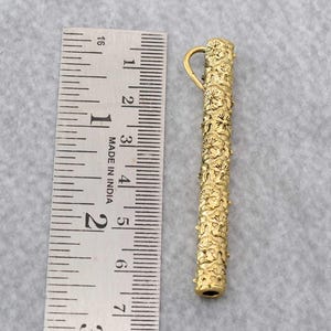 Puede incluir: Un colgante dorado con un diseño floral, de aproximadamente 9 cm de largo. El colgante tiene forma cilíndrica con un lazo en la parte superior. El patrón floral cubre toda la superficie del colgante.