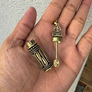 May include: Two antique brass items are shown. One is a cylindrical container with detailed rope and geometric patterns. The other is a small spoon-like tool with a decorative handle. Both items have a vintage appearance.