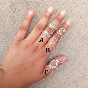 Puede incluir: Tres anillos de plata, etiquetados como A, B y C, se llevan en los dedos de una persona. El anillo A es una pila de tres bandas finas, el anillo B es una sola banda con un diseño curvo y el anillo C es una banda gruesa con un diseño más intrincado.