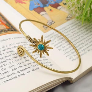 Può includere: Un braccialetto dorato a forma di sole con una pietra centrale turchese. Il braccialetto ha un design a spirale su un'estremità ed è appoggiato su un libro aperto con testo.