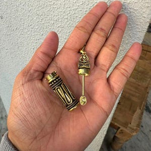Könnte beinhalten: Ein goldfarbener Metallanhänger bestehend aus einem zylindrischen, texturierten Gehäuse mit kompliziertem Muster und einem passenden abnehmbaren Deckel, der an einem dünnen Stab mit einem kleinen, ovalen Löffel am Ende befestigt ist.