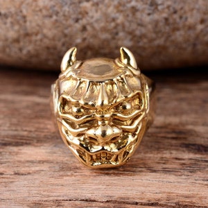 May include: A gold-coloured ring featuring a detailed design of a demon's face. The ring has two horns on top and a menacing expression.