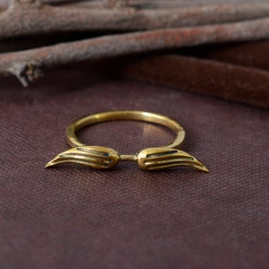 May include: A gold-coloured ring with a wing design. The ring features two stylised wings extending outwards from the band. The wings have detailed feather-like textures. The ring is set against a dark, textured background.