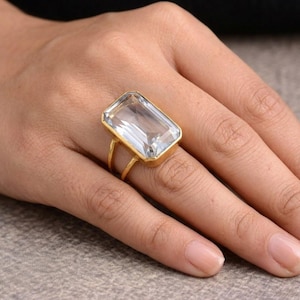 Op de afbeelding: Een goudkleurige ring met een grote, rechthoekige, heldere edelsteen. De ring heeft een eenvoudige, gladde band en de edelsteen is gezet in een gouden bezel. De ring wordt aan een vinger gedragen en toont zijn grootte en helderheid.