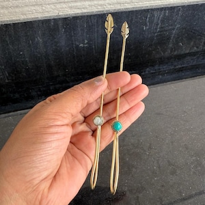 Puede incluir: Dos palitos de pelo dorados con puntas en forma de pluma. Un palito tiene una piedra blanca en la parte inferior, y el otro tiene una piedra turquesa.