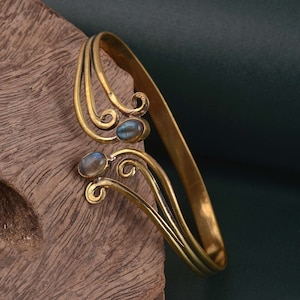 Könnte beinhalten: Ein goldfarbenes Armband mit einem filigranen, verzierten Design mit zwei blaugrünen Edelsteinen. Das Armband ist offen und verstellbar.