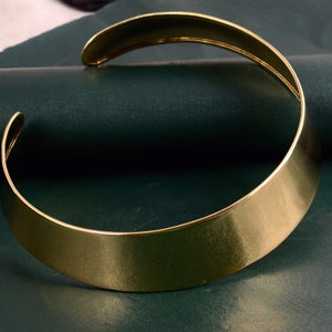 Może przedstawiać: Szeroki złoty naszyjnik choker. Naszyjnik jest płaski i ma gładkie, polerowane wykończenie.