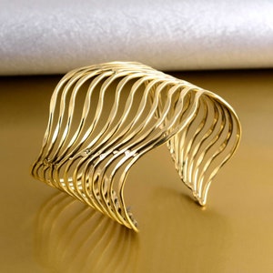Peut inclure: Un bracelet manchette en métal doré avec un design ondulé et superposé. Le bracelet est ouvert et a une forme large et incurvée.