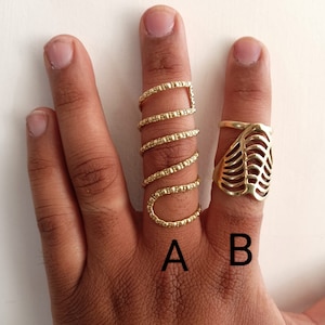 May include: Three gold rings on a hand. The ring on the index finger is a simple band with a textured surface. The ring on the middle finger is a set of three bands with a textured surface. The ring on the ring finger is a leaf-shaped design.