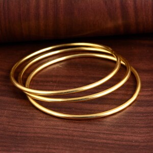 May include: Three gold bangles stacked on top of each other on a brown wooden surface.