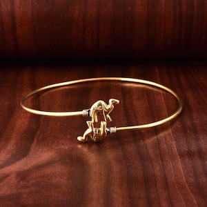 May include: A gold-coloured bangle bracelet with two camel figures. The bracelet has a simple, open design with the camels at the ends. The camels have visible humps and legs. The bracelet is displayed on a dark brown wooden surface.