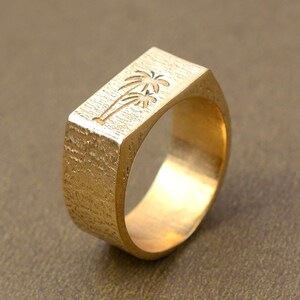 Puede incluir: Un anillo de color dorado con una banda de forma cuadrada que presenta un diseño de palmera grabado.