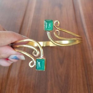 Könnte beinhalten: Ein goldfarbenes Armreif-Armband mit zwei grünen Edelstein-Akzenten. Das Armband hat ein zartes, verziertes Design.