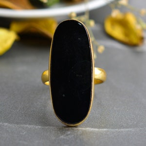 Puede incluir: Un anillo dorado con una gran piedra negra de forma ovalada. La banda y el engaste del anillo tienen un acabado metálico pulido. La piedra negra es lisa y brillante, creando un contraste con el dorado.