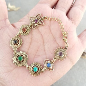 May include: Gold bracelet with seven charms, each featuring a different colored gemstone. The charms are connected by a gold chain.
