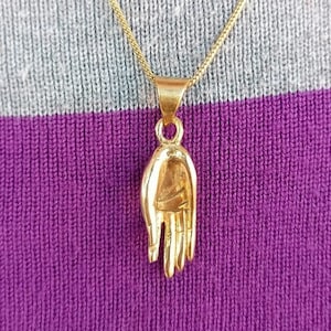Op de afbeelding: Een gouden ketting met een gedetailleerd handvormig hanger. De hanger hangt aan een dunne gouden ketting.