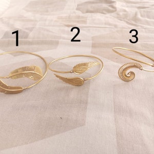 Pode incluir: Três pulseiras de metal dourado com designs diferentes. A primeira pulseira apresenta dois pingentes em forma de pena. A segunda pulseira apresenta um único pingente em forma de asa. A terceira pulseira apresenta um pingente em forma de espiral.