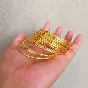 Pode incluir: Uma pilha de pulseiras douradas, provavelmente de metal, são seguradas em uma mão. As pulseiras são lisas e brilhantes.