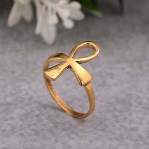 Könnte beinhalten: Ein goldfarbener Ring mit einem Ankh-Symbol-Design. Der Ring ist aus Metall und hat eine glatte Oberfläche.