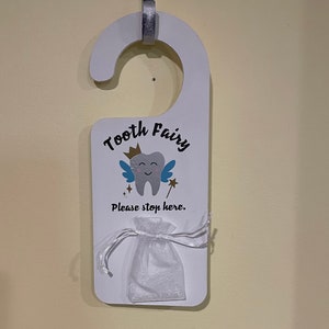 May include: White wooden door hanger with a tooth fairy design. It features a blue tooth with wings and a gold crown. The text "Tooth Fairy" is written in black and the text "Please stop here." is written in black below the tooth. A white mesh bag is hanging from the bottom of the hanger.