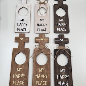 May include: Wooden door hangers in various shades of brown, white, and beige. Each hanger features a cross-shaped top with a heart detail and a circular cutout. The phrase "MY HAPPY PLACE" is printed on each hanger.