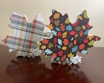 May include: Two wooden maple leaf decorations, one with a plaid pattern and the other with a pattern of colorful leaves on a dark brown background. Both leaves have white twine bows.
