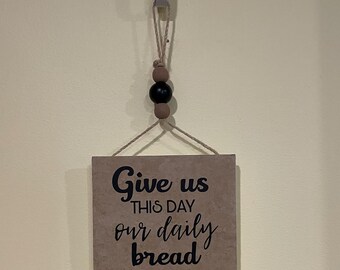 May include: A decorative wall hanging with the text "Give us this day our daily bread." The sign is brown with black lettering and a decorative leaf design. It hangs from a metal hook with a rope and wooden beads.