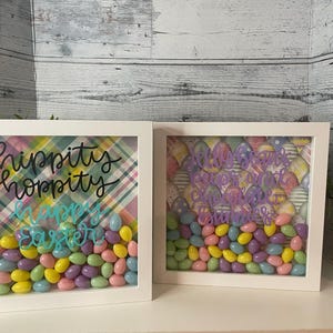 May include: Two white shadow boxes with a plaid and floral patterned background. The boxes are filled with pastel-coloured candy eggs and have the text "Hippity Hoppity Happy Easter" written in black and turquoise.