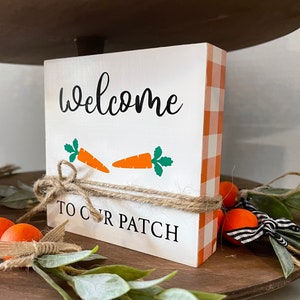 May include: White wooden block with orange and white gingham fabric wrapped around it. The block has the text "Welcome to our patch" written on it in black. There are two orange carrot cutouts on the block.