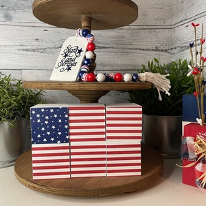 Peut inclure: Un plateau en bois à plusieurs niveaux décoré d'objets patriotiques rouge, blanc et bleu. Le niveau supérieur présente une pancarte en bois sur laquelle est inscrit "Stars and Stripes" et une guirlande de perles. Le niveau intermédiaire est composé de quatre blocs de bois empilés pour former un drapeau américain. Le niveau inférieur présente un ruban à carreaux rouge et blanc noué autour d'un bloc de bois avec un dessus blanc.