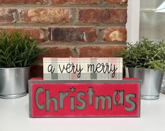 May include: A Christmas-themed wooden block sign with the words "Christmas" cut out in green against a red background. A smaller block above reads "a very merry" in script. Greenery in silver pots flank the sign.