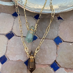 Pode incluir: Um colar em camadas com duas correntes de metal dourado com elos ovais alongados. Cada corrente sustenta um pingente em forma de pequena garrafa de cristal facetado com tampa de metal dourado. Uma garrafa &eacute; verde-clara e a outra &eacute; castanho-escura.
