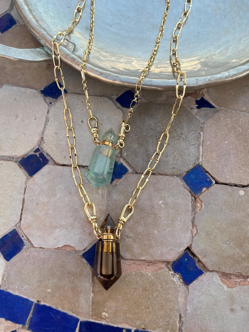Pode incluir: Um colar em camadas com duas correntes de metal dourado com elos ovais alongados. Cada corrente sustenta um pingente em forma de pequena garrafa de cristal facetado com tampa de metal dourado. Uma garrafa &eacute; verde-clara e a outra &eacute; castanho-escura.