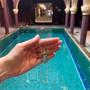 Pode incluir: Um colar de corrente dourada com um pequeno pendente de frasco de perfume de cristal transl&uacute;cido em verde p&aacute;lido. O pendente tem uma forma hexagonal facetada com uma tampa de metal dourado e est&aacute; suspenso numa delicada corrente de elos.