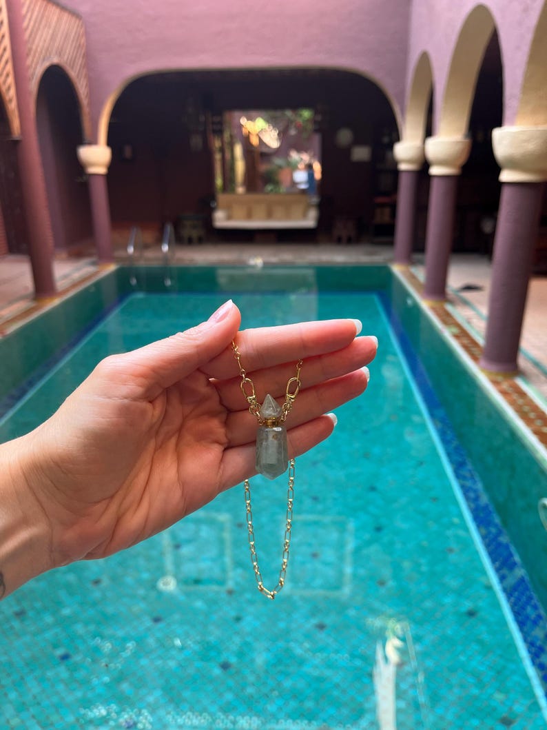 Pode incluir: Um colar de corrente dourada com um pequeno pendente de frasco de perfume de cristal transl&uacute;cido em verde p&aacute;lido. O pendente tem uma forma hexagonal facetada com uma tampa de metal dourado e est&aacute; suspenso numa delicada corrente de elos.