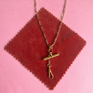 Könnte beinhalten: Eine goldfarbene Uhrenkette mit T-Bar und Federverschluss, präsentiert auf einem roten, rautenförmigen Stoff vor rosa Hintergrund. Die Kette besteht aus kleinen, rechteckigen Gliedern.