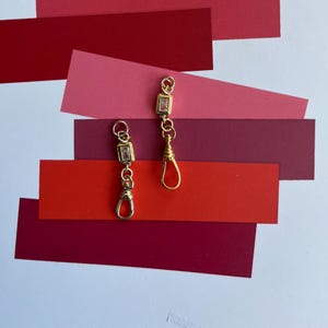 May include: Two gold-coloured earrings, each with a rectangular clear stone and decorative chain. The earrings have a clasp at the bottom. The background is composed of layered red and pink paper in various shades.