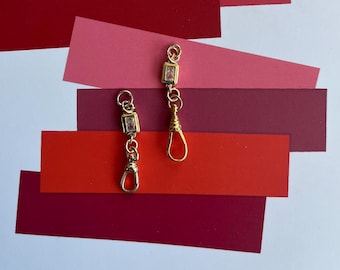 May include: Two gold-colored, rectangular-shaped earrings with a small, clear stone in the center of each. The earrings have a decorative chain and a clasp at the bottom. The background features layered red and pink paper.
