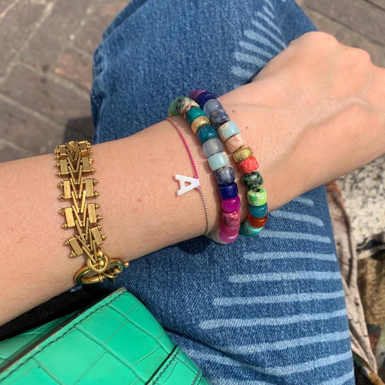 May include: A gold chain bracelet with a clasp, a pink string bracelet with a white letter A charm, and a multi-colored beaded bracelet with a variety of stones.