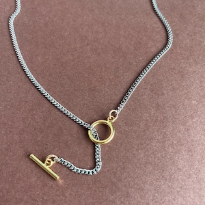 May include: A silver chain necklace with a gold-colored circle pendant and a gold bar closure. The necklace is displayed on a brown surface. The jewelry is a simple, elegant design.