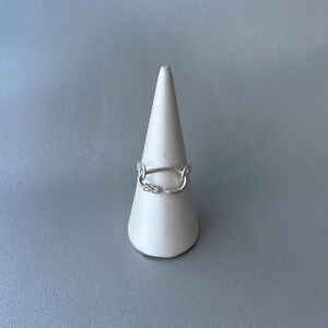 以下が含まれることがあります： ユニークなデザインのシルバーリング。白い円錐形のスタンドに展示されています。リングは、装飾的なディテールが施されたオープンな楕円形をしています。リングは光沢のある金属素材で作られています。