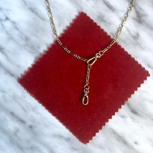 May include: A fine gold-coloured necklace with a chain design and a clasp. The necklace is displayed on a red square fabric, set against a white and grey marble surface. The necklace has a small, dangling charm.