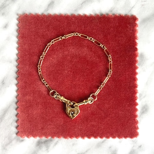 May include: A gold-colored bracelet with a heart-shaped lock pendant. The bracelet is on a red velvet surface. The heart-shaped lock has intricate detailing. The bracelet's chain is made of small, rectangular links.