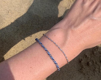 May include: Two bracelets on a wrist. One bracelet is a thin gold chain, the other is a thicker braided cord with blue and white threads.
