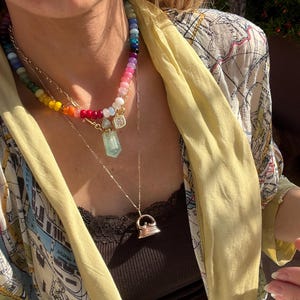 Pode incluir: Um conjunto de colares em camadas com um fio de contas multicoloridas em gradiente arco-&iacute;ris, uma corrente dourada com um pingente de frasco de perfume de cristal transparente e uma segunda corrente dourada com um berloque de ferro de passar.