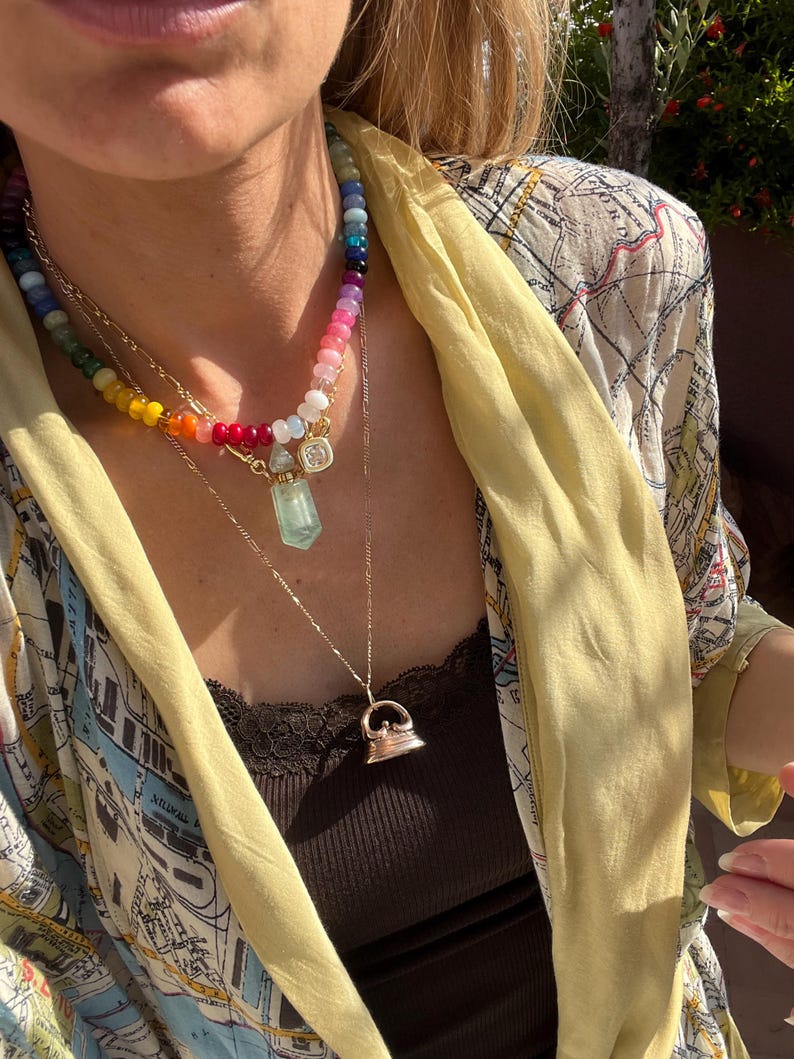 Pode incluir: Um conjunto de colares em camadas com um fio de contas multicoloridas em gradiente arco-&iacute;ris, uma corrente dourada com um pingente de frasco de perfume de cristal transparente e uma segunda corrente dourada com um berloque de ferro de passar.