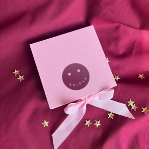 Pode incluir: Uma caixa de presente quadrada rosa claro amarrada com uma fita rosa claro correspondente. O centro da caixa apresenta um adesivo circular roxo escuro com dois pontos brancos e o texto BOIDAE impresso em branco. Pequenas lantejoulas douradas.