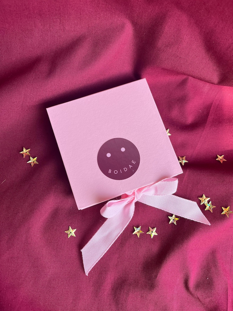 Pode incluir: Uma caixa de presente quadrada rosa claro amarrada com uma fita rosa claro correspondente. O centro da caixa apresenta um adesivo circular roxo escuro com dois pontos brancos e o texto BOIDAE impresso em branco. Pequenas lantejoulas douradas.